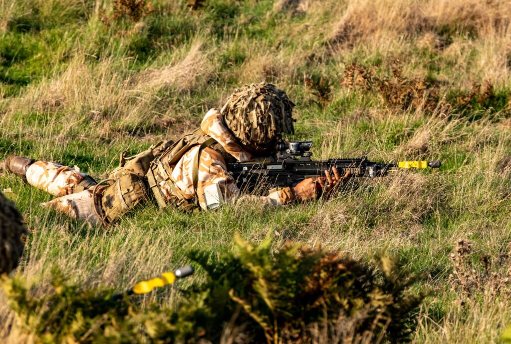 British soldier in camouflage and helmet net, lying prone in long grass aiming a rifle with a yellow training attachment during a field exercise