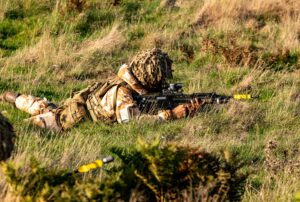 British soldier in camouflage and helmet net, lying prone in long grass aiming a rifle with a yellow training attachment during a field exercise